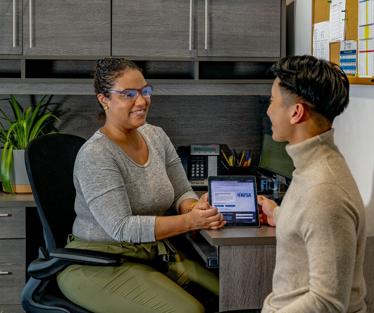 Admissions advisor discussing financial aid with a prospective student in an office, reviewing FAFSA information on a tablet.
